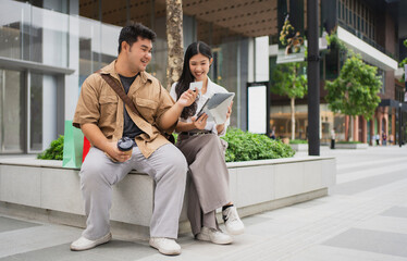 Two Asian a while walking outdoors at a large-scale retail complex. They are holding various shopping bags, suggesting a successful day of purchasing goods together