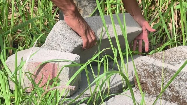Close-up of a worker's hands lifting Autoclaved Aerated Concrete (AAC) blocks from the ground. Set in an outdoor construction area, showing manual labor, building materials, and masonry work