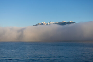 Snowy Mountains Above Sea Fog.Snow-covered mountain peaks rising above low coastal fog over calm blue sea under clear sky. Serene winter landscape with mist, horizon, and minimal composition.