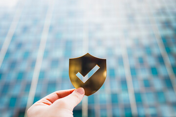 Hand holding a golden shield icon with check mark symbol on blur modern glass building background, representing cybersecurity, trust, data protection, online privacy, and secure technology concept.