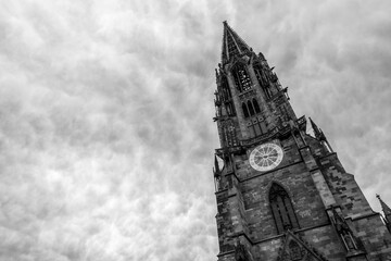 Dramatic black and white view of a historic cathedral tower with clock, rising into a cloudy sky. Timeless fine art architecture scene with strong perspective, gothic details, and moody atmosphere