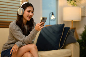 Obraz premium A young Asian woman relaxes on a sofa wearing headphones, smiling while listening to music and podcasts on her smartphone at home