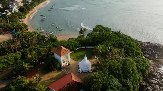 Aerial drone footage of Unawatuna Beach in Sri Lanka with traditional fishing boats on the shore and a Buddhist temple near the coastline. Tropical ocean, sandy beach and cultural landmarks.