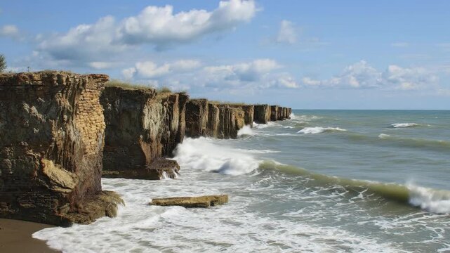 Dynamic wide shot capturing powerful ocean waves crashing against rugged, layered cliffs under a clear blue sky dotted with fluffy white clouds. The intense force of the sea creates dramatic white foa