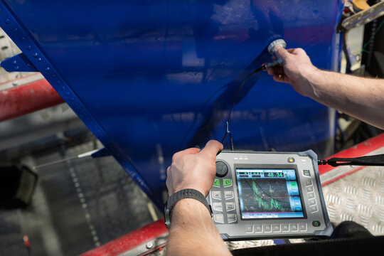 An aviation technician conducting ultrasonic testing on a blue aircraft rudder using portable NDT equipment with a digital display for defect detection, structural integrity inspection and maintenance