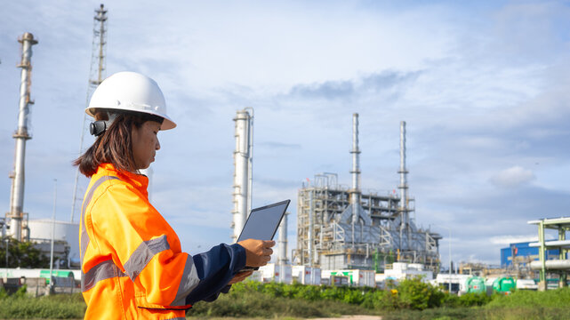A worker stands outside an industrial plant, holding a tablet. The facility has tall structures and pipes visible behind. The sky is clear and shows daylight - Powered by Adobe