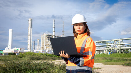 A worker in an orange safety jacket and hard hat stands outside a processing plant. She uses a laptop to check data and monitor tasks near tall structures and machinery