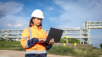 A worker stands at an industrial location with a laptop, engaged in her tasks. She wears safety gear, including a hard hat and reflective clothing