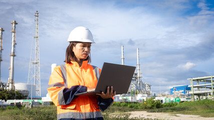 A worker in safety gear stands outside on an industrial site. She uses a laptop to check information. Tall structures and equipment are visible in the background. The sky is partly cloudy