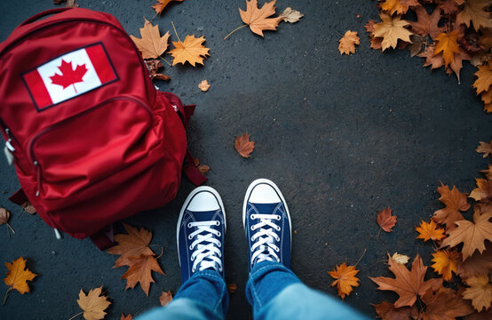 Red backpack with Canadian flag patch and blue sneakers on asphalt road surrounded by autumn leaves. Represents Canadian journey or study abroad experience. Travel concept.