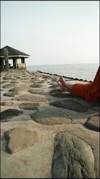 Person wearing orange pants relaxing on a rocky beach with a gazebo in the background under a clear sky.
