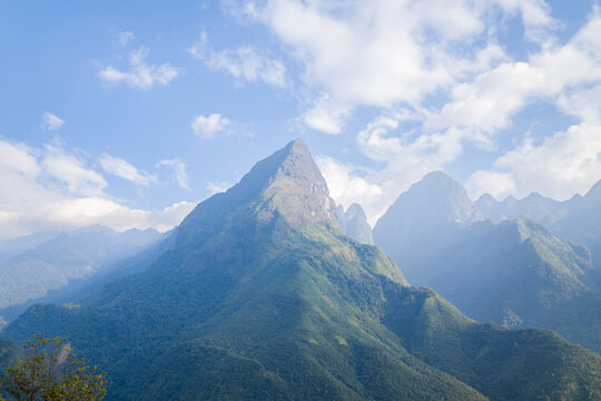 A dramatic green mountain rises sharply beneath a partly cloudy sky, with misty ridges and lush forest covering the slopes in the Fansipan massif of northern Vietnam.