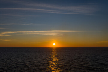 Fototapeta premium Warm golden sunlight reflects across calm open water as the sun sets above the horizon, creating a tranquil seascape between the Adriatic and Ionian Seas. Wispy clouds and a gradient sky enhance the