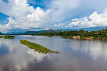 Obraz premium Large cumulus clouds tower above the Mekong River as it winds past grassy islets and tropical banks near Luang Prabang, Laos. Wooden boats line the lush shoreline, with distant mountains providing a