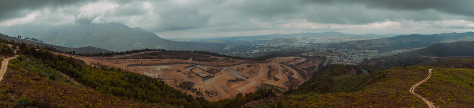 A panoramic view overlooking Stellenbosch, South Africa, showing the valley floor and surrounding mountains under a heavy, moody sky. The foreground reveals rugged hillside trails and forestry.
