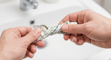 Man squeezing the very last bit of toothpaste out of a crumpled metal tube, illustrating resourcefulness and household economy.