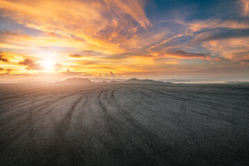 Empty asphalt road with tire marks under a dramatic sunset sky with mountains in the background. © zhao dongfang