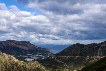 Fototapeta premium Expansive view over a rugged mountain valley descending toward the blue sea, with winding roads and scattered greenery beneath a sky filled with textured, dramatic clouds. The landscape blends rocky