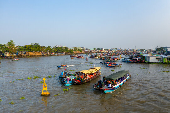 Tourist boats with covered roofs move along the busy river at Cai Rang floating market in Can Tho, Mekong Delta, Vietnam. The wide view shows numerous vessels, riverside houses, and greenery under