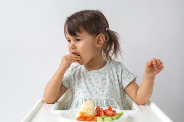 A little girl sitting in a high chair and eating fresh vegetables. She is focused while putting a piece of food in her mouth. Concept of healthy eating and childhood development