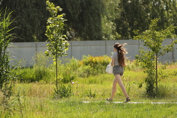 A young girl in a revealing outfit with bare legs walks along a park alley. A walk in the fresh air among summer greenery. Fashion of modern youth.
