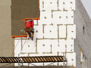 A builder in climbing equipment insulates the facade of a high-rise building with foam plastic. Safety measures for high-altitude construction work. Heat conservation in homes and energy savings