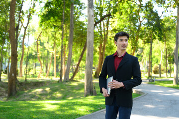 A man walks through a green park holding a laptop and a coffee cup on a bright day © Prathankarnpap
