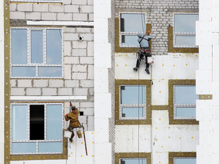A builder in climbing equipment insulates the facade of a high-rise building with foam plastic. Safety measures for high-altitude construction work. Heat conservation in homes and energy savings