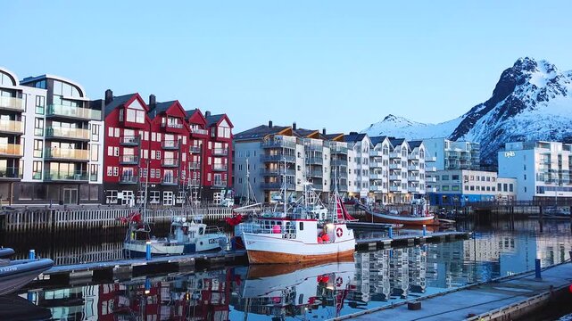 Footsteps echo along the harbor as morning light shines on red and gray buildings. The water is still, showing clear reflections of the structures. Boats are anchored nearby.