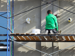 A worker plastering a building facade while standing on a scaffold. Construction work on the construction of a high-rise building. Safety measures for high-rise construction works. Insulation of thin