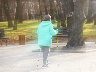 Nordic walking. An elderly woman in urban clothes is walking in the park in the rays of sunlight. Healthy lifestyle. Sporting event. Activity of the elderly. Wellness exercises in the fresh air
