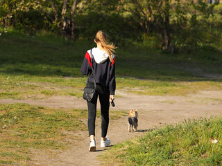 A young girl walks in the park with a dog. A sporty blonde walks a pet in nature. View of a beautiful girl from the back. Relax in the fresh air with your loved one. Friendship between children