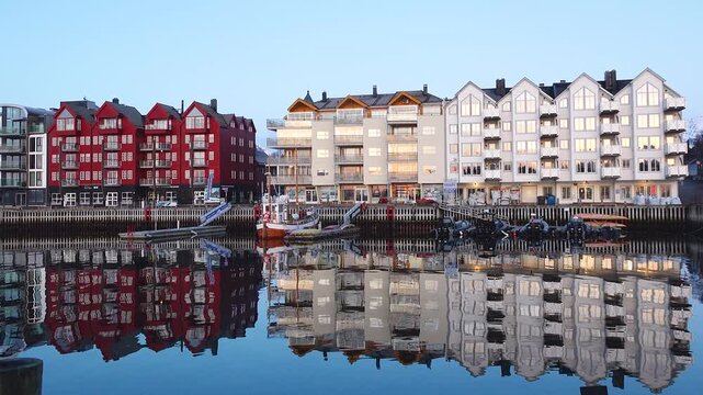 Footsteps echo along the harbor as morning light shines on red and gray buildings. The water is still, showing clear reflections of the structures. Boats are anchored nearby.