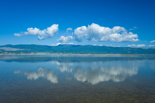 White cumulus clouds are reflected on the calm surface of the Missolonghi salt marsh in western Greece. The expansive wetland features distant mountains, clear blue sky, and tranquil water creating a