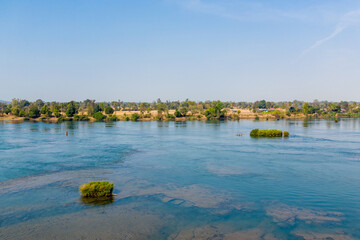 Fototapeta premium Wide stretch of the Mekong River flows past small green islets and a tree-lined riverbank in the 4000 Islands region of southern Laos. Crisp daylight highlights the calm blue water and scattered