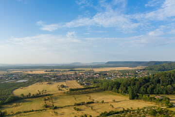 Fototapeta premium Expansive aerial view of Hattonchatel village surrounded by golden farmland, orchards, and dense green woodland under a bright blue sky. The composition highlights the patchwork fields, rural