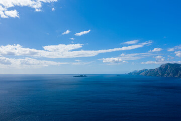 Fototapeta premium Wide expanse of deep blue sea with small islands on the horizon and rugged coastal mountains to the right, all beneath a sky filled with scattered white clouds. The scene is illuminated by bright