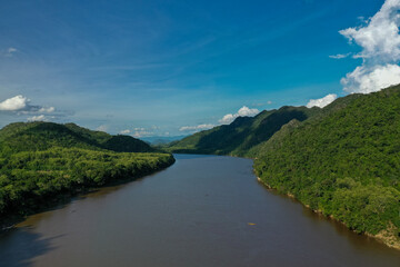 Fototapeta premium A broad stretch of the Mekong river flows between densely forested green hills under a vivid blue sky with scattered clouds. The scene features rich tropical vegetation, gentle slopes, and tranquil