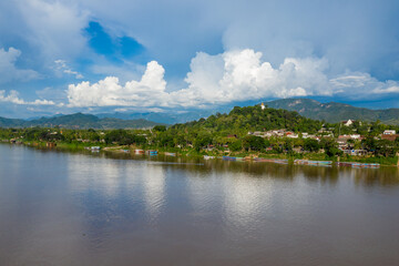Fototapeta premium A golden stupa crowns a lush green hill overlooking the Mekong River in Luang Prabang, Laos. Colorful boats line the riverbank beneath dramatic cumulus clouds and distant blue mountains.
