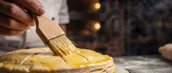 Professional baker brushing egg wash on a raw Galette des Rois. Preparation of traditional French Epiphany cake in a bakery. Close-up of artisan hand glazing the puff pastry.