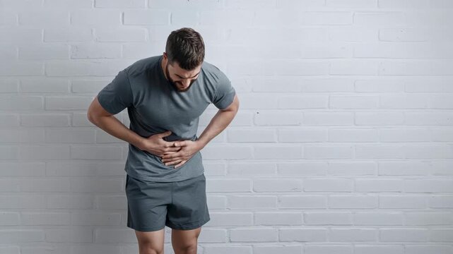 Adult man bending forward and holding his stomach while standing against a plain indoor wall