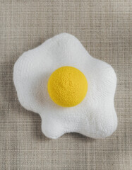 Cute handmade fried egg made of white and yellow wool felt. whimsical and simple craft object on textured fabric, playful imitation of breakfast food evoking charming mood