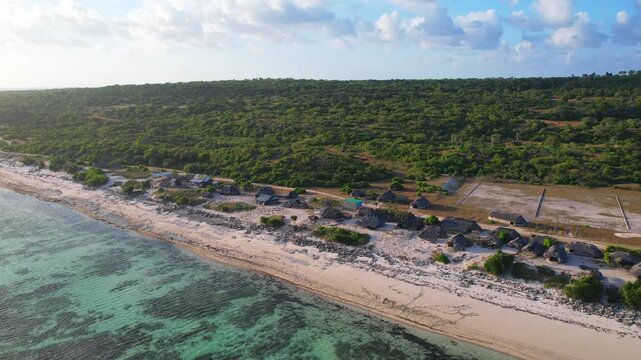 Aerial drone footage of many seaweed wooden drying thatch house farms, with white sand beach, a transparent blue sea water with coral reef coastal area, a forest, at sunset, in Sabu island, Indonesia