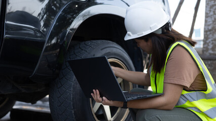 A woman in a safety vest and hard hat inspects a vehicle's tire while using a laptop. She focuses on her task in a parking area under bright sunlight