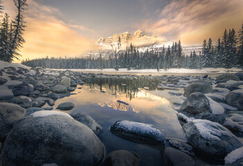 View of snow-dusted rocks leading to the serene waters reflecting the snow-capped mountain and evergreen trees under a sky painted with warm hues, Banff, Alberta, Canada.