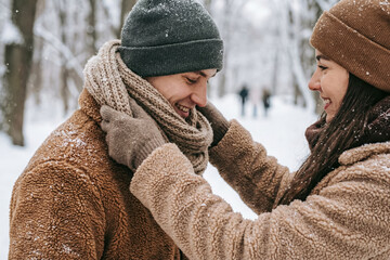 Young couple adjusting scarf outdoors in winter forest, warm clothing and tender care during snowfall, love and seasonal connection, generative AI