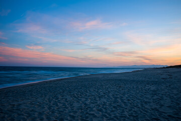 Obraz premium Soft pink and orange hues illuminate the sky above the empty sandy beach at Plage de Acchio, Fiumicello. Gentle waves and distant mountains create a peaceful, expansive seascape in the fading evening