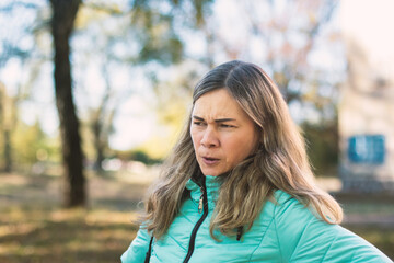 A pensive middle-aged woman with a concerned expression, wearing a teal jacket, stands in a park. Her furrowed brow suggests worry or deep thought in natural light.