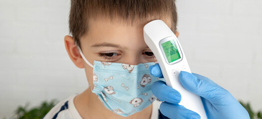 Female doctor in face mask measuring boy's temperature with infrared thermometer
