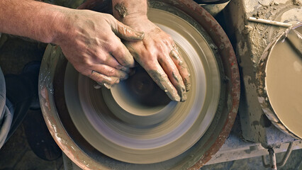 A close-up of a craftsperson's hands shaping clay on a pottery wheel, creating a spinning mound of earth in a workshop filled with tools and clay remnants.